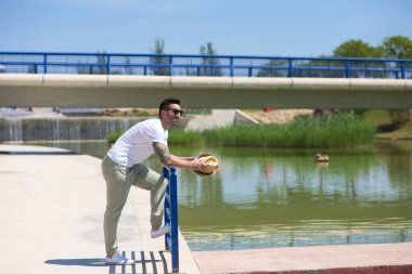Parktaki gölün kenarındaki korkuluklara yaslanmış günlük kıyafetler ve güneş gözlüğü takan yakışıklı genç adam elinde bir gölge tutuyor ve tatilin tadını çıkarıyor.