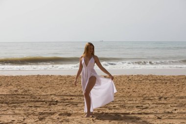 Beyaz elbiseli güzel sarışın kadın güneşli bir günde kumsalın kıyısında yürüyor. Kadın farklı vücut ifadeleri kullanıyor. Arka planda mavi deniz.