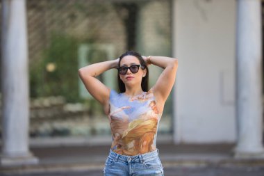 Uzun saçlı ve güneş gözlüklü güzel esmer kadın ellerini başının arkasına koyuyor, kadın gündelik ve modern kıyafetler giyiyor.