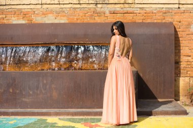 Uzun kıvırcık saçlı güzel İspanyol kadın, çiçekli ve parlak taşlı somon rengi bir elbise giymiş bir model gibi poz veriyor. Kadın demir bir çeşmenin önünde duruyor.