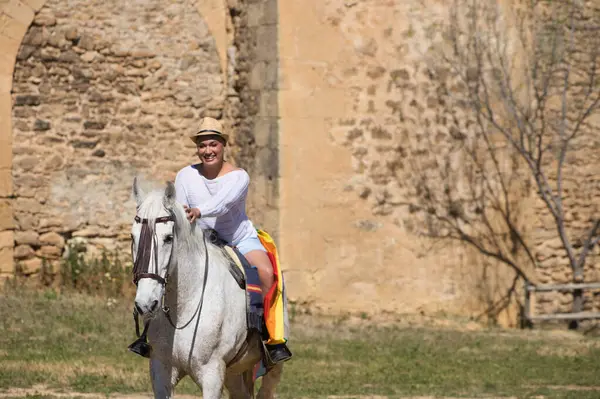 Young non-binary latin person, riding a white horse with the gay pride flag. In the background a medieval castle. Concept of diversity, homosexuality and human rights