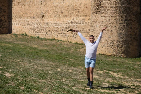 Portrait of young non-binary latin person walking with a straw hat in his hand while having fun. In the background a medieval castle. Concept of diversity, homosexuality and human rights