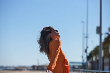 Turuncu elbiseli güzel sarışın bir kadının profil portresi plajda ahşap korkuluklara yaslanıyor. Ufukta mavi gökyüzü görünüyor. Moda ve güzellik konsepti