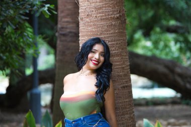 Genç ve güzel esmer kadın ve Latin, yeşillik ve yeşillik ile kaplı bir parkta bir hurma ağacının gövdesine yaslanmış. Kadın gülümsüyor ve mutlu. Mutluluk kavramı. Sarı gün, 20 Haziran