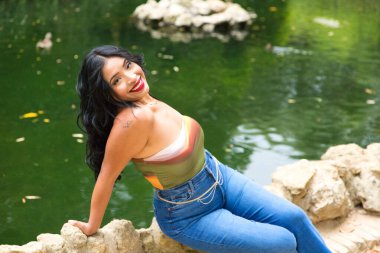 Genç ve güzel esmer kadın ve Latin, ördeğin taştan kenarında ve parktaki balık havuzunda oturuyor. Kadın gülümser, mutludur ve farklı ifadeler kullanır.