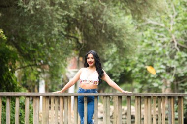 Genç ve güzel esmer kadın ve Latin, ördeğin üzerindeki ahşap köprünün korkuluğuna yaslanıyor ve parktaki balık göletine yaslanıyor. Kadın gülümsüyor, mutlu ve günün tadını çıkarıyor..