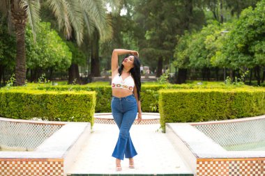 Güney Amerikalı genç ve güzel bir Latin kadın parkın ağaçlarının ve bitkilerinin arasında yürüyor. Kadın gülümsüyor, mutlu ve fotoğraf için poz veriyor. Seyahat ve tatil konsepti