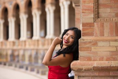 Kısa kırmızı elbiseler ve kırmızı ayakkabılar giymiş genç ve güzel esmer ve Latin bir kadın İspanya 'nın Seville Meydanı' nı ziyaret ediyor. Kadın tuğla bir sütuna yaslanıyor ve mutlu. Seyahat kavramı