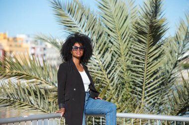 Afro saçlı, genç ve güzel bir kadın. Günlük giysiler içinde. Seville, İspanya 'da tatilinin tadını çıkarırken korkuluklara yaslanıyor. Arka planda Seville nehri.