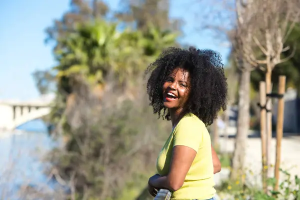 Young, pretty black woman with afro hair and sunglasses wearing jeans and yellow shirt leans on the railing. In the background the river guadalquivir and the neighbourhood in seville.