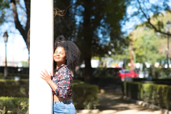 Young, beautiful black woman with afro hair dressed in casual clothes leaning against a marble column in the park. The woman enjoys the sunny day while making different expressions
