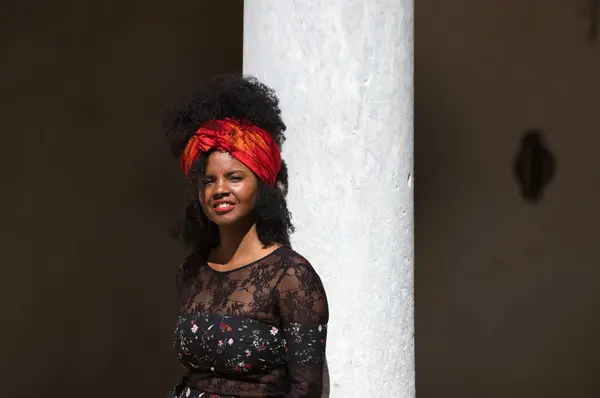 Portrait of a young, beautiful black woman with afro hair and black dress with flowers, wearing a red scarf in her hair. The woman is happy and laughing and having fun leaning on a marble column