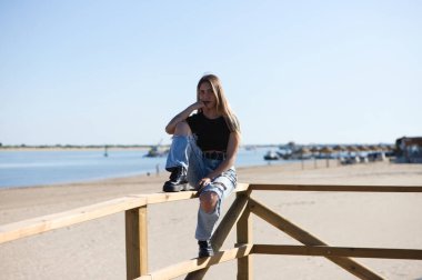 Yırtık kotlu, siyah bluzlu ve çizmeli güzel bir kız. Kız asi ve sert bir karakteri var. Kız plaj korkuluklarında oturuyor.