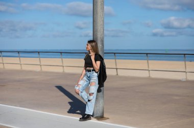 Genç, güzel sarışın kadın sokak lambasına yaslanıyor. Kadın gündelik kıyafetler giyiyor ve omzunun üzerine siyah deri bir ceket giyiyor. Arka planda deniz ve mavi gökyüzü