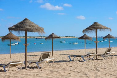 Güneşli bir günde kumsalda saman şemsiyeler ve güneş şezlongları. Dinlenme, rahatlama, tatil ve seyahat kavramı. Arka planda balıkçı tekneleri var.