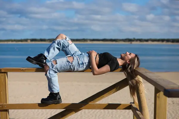 Pretty young girl in ripped jeans and black top and boots. The girl is rebellious and with a tough character. The girl is lying on the railing of the beach promenade