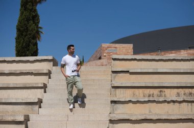 Günlük kıyafetler ve güneş gözlüğü takmış yakışıklı genç adam tribünün merdivenlerinden aşağı iniyor ve mekanın farklı yerlerine bakıyor.