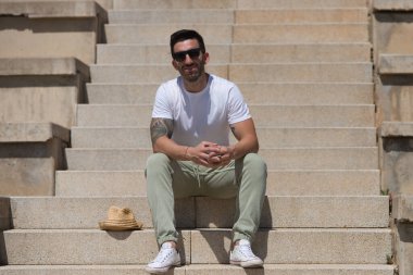 Parktaki merdivenlerde oturan, günlük kıyafetler ve güneş gözlüğü takan yakışıklı genç adam. Adamın bir şapkası var ve baharın güneşli gününün tadını çıkarıyor. Seyahat ve tatil konsepti