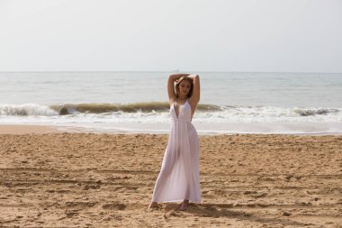 Beyaz elbiseli güzel sarışın kadın güneşli bir günde kumsalın kıyısında yürüyor. Kadın farklı vücut ifadeleri kullanıyor. Arka planda mavi deniz.