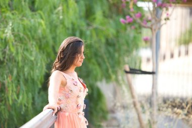 Uzun kıvırcık saçlı güzel Latin kadın, çiçekli ve parlak taşlı zarif bir elbise giymiş bir model gibi poz veriyor. Arka planda nehir var. Kadın bir bacağını açıkta bırakıyor.