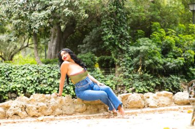 Genç ve güzel esmer kadın ve Latin, ördeğin taştan kenarında ve parktaki balık havuzunda oturuyor. Kadın gülümser, mutludur ve farklı ifadeler kullanır.