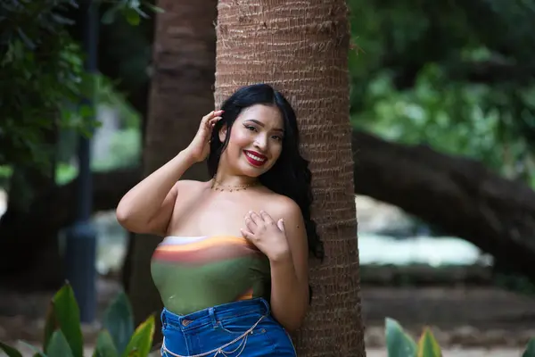 Genç ve güzel esmer kadın ve Latin, yeşillik ve yeşillik ile kaplı bir parkta bir hurma ağacının gövdesine yaslanmış. Kadın gülümsüyor ve mutlu. Mutluluk kavramı. Sarı gün, 20 Haziran