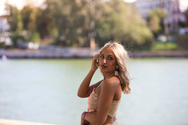 Young beautiful blonde woman dressed in embroidered trousers and crochet top by the river bank in seville. In the background a mediterranean style neighbourhood with white painted houses