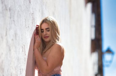 Genç, güzel, mavi gözlü sarışın kadın duvara yaslanıyor ve gündelik, modern kıyafetler giyiyor. Kadın yüzüyle farklı ifadeler kullanıyor, gülümsüyor, ciddi, mutlu ve üzgün.