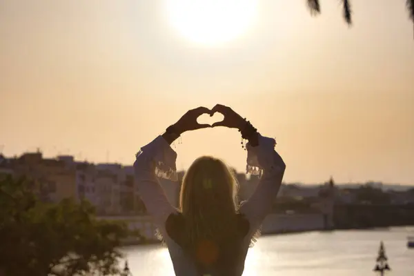Beautiful young blonde girl from the USA makes a heart with her hands with her arms raised to the sky. Rays of sunlight pierce the heart. It is the golden hour
