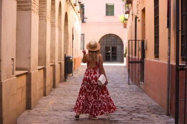 Genç ve güzel sarışın kadın, güneş gözlüğü ve kafasında hasır bir şapkayla Seville 'in tarihi merkezinde yürüyor. Kadın tatilde ve şehrin tadını çıkarıyor. Kadın mutlu.