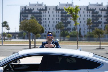 Sakallı ve mavi takım elbiseli yakışıklı genç iş adamı beyaz spor arabasına yaslanıyor. Adam güneş gözlüğü takıyor. İş seyahati kavramı
