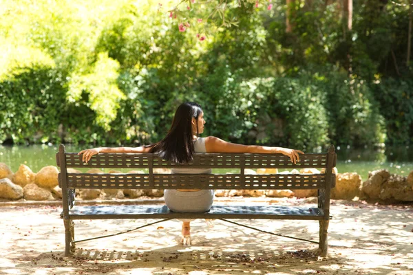 Latin woman, young and beautiful brunette relaxed and sitting on a bench looking at the pond and trees in the area. The photo is taken from behind