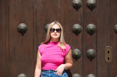 Genç, güzel sarışın kadın ahşap bir kapının önünde. Kadın modern günlük kıyafetler ve güneş gözlüğü takıyor ve farklı vücut duruşları ve ifadeleri sergiliyor.