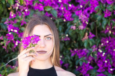 Siyah giyinmiş genç, güzel sarışın kadın bir gözünü mor bir bougainvillea çiçeğiyle kaplıyor ve önüne bakıyor. Arka planda dev bir begonvil bitkisi var.