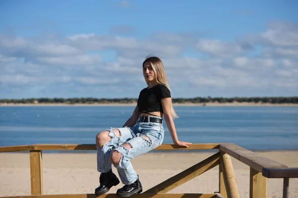 Pretty young girl in ripped jeans and black top and boots. The girl is rebellious and with a tough character. The girl is sitting on the railing of the beach promenade