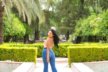 Genç ve güzel esmer bir kadın, Latin kökenli, parkın ağaçlarının ve bitkilerinin arasında yürüyor. Kadın gülümsüyor ve mutlu ve fotoğraflara arkasını dönmüş poz veriyor..