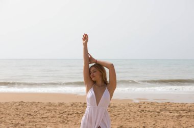 Beyaz elbiseli güzel sarışın kadın güneşli bir günde kumsalın kıyısında yürüyor. Kadın farklı vücut ifadeleri kullanıyor. Arka planda mavi deniz.