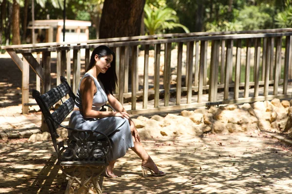 Latin woman, young and beautiful brunette relaxed and sitting on a bench looking at the pond and trees in the area. The woman is wearing an elegant grey dress.