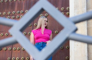 Genç, güzel, mavi gözlü sarışın kadın ahşap bir kapının önünde duruyor ve gündelik, modern kıyafetler giyiyor. Kadın elmas şekilli ızgaradan kameraya bakıyor.