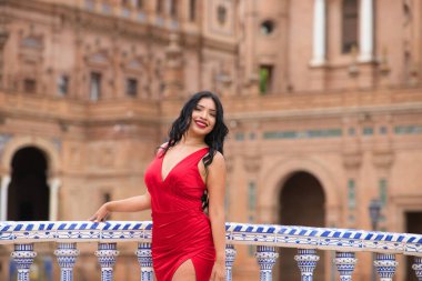 Kısa kırmızı elbiseler ve kırmızı ayakkabılar giymiş genç ve güzel esmer ve Latin bir kadın İspanya 'nın Seville Meydanı' nı ziyaret ediyor. Bir kadın fayanslı bir köprünün parmaklıklarına yaslanıyor ve mutlu..