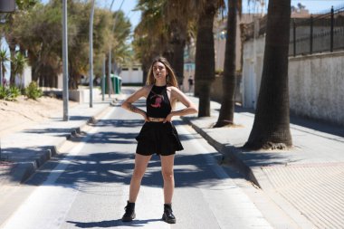 Siyah etek ve siyah bluz giymiş sarışın, genç ve güzel bir kadın yaya geçidinden geçiyor. Kadın karşıdan karşıya geçiyor ve farklı pozlar verme fırsatını yakalıyor.