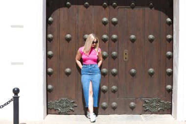 Genç, güzel sarışın kadın ahşap bir kapının önünde. Kadın modern günlük kıyafetler ve güneş gözlüğü takıyor ve farklı vücut duruşları ve ifadeleri sergiliyor.