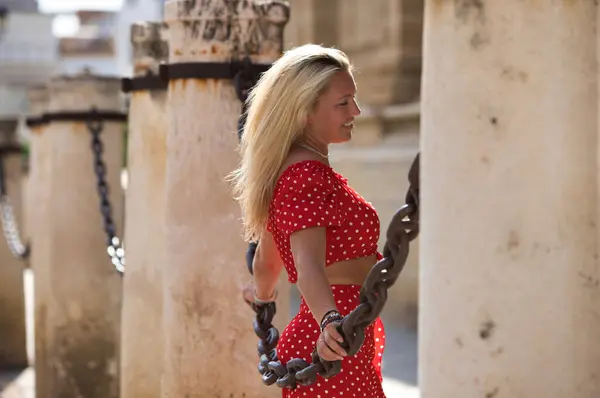 Beautiful young blonde woman from the United States is on a sightseeing trip in Seville, Spain. The girl leans between the columns and chains surrounding the city\'s gothic-style cathedral