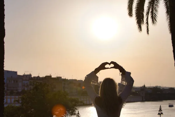 Beautiful young blonde girl from the USA makes a heart with her hands with her arms raised to the sky. Rays of sunlight pierce the heart. It is the golden hour