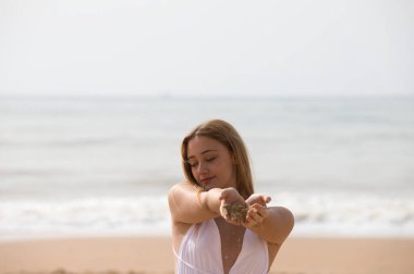 Beyaz elbiseli güzel sarışın kadın ellerini önündeyken ellerinin arasından kum döküyor. Kadın mutlu ve eğleniyor. Arka planda mavi deniz.