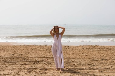 Beyaz elbiseli güzel sarışın kadın güneşli bir günde kumsalın kıyısında yürüyor. Kadın farklı vücut ifadeleri kullanıyor. Arka planda mavi deniz.