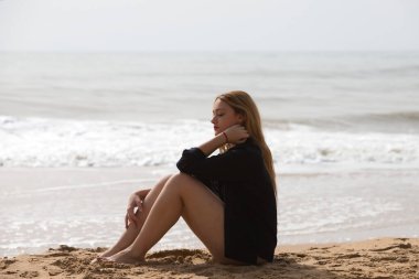 Siyah gömlekli güzel sarışın kadın sahilde üzgün ve depresif bir şekilde oturuyor. Kadın dalgın ve yere bakıyor bitkin ve yorgun. Kadın acı çekiyor..