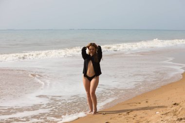 Genç ve güzel sarışın kadın siyah gömlek ve siyah bikini giyip sahilde yürüyor ve farklı ifadeler ve duruşlar sergiliyor. Arka planda deniz ve gökyüzü.