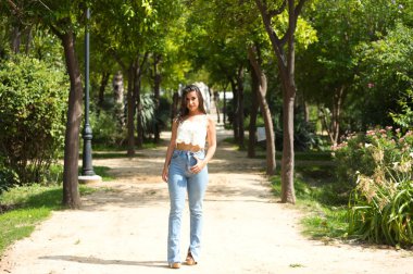 Uzun kıvırcık saçlı, güzel, koyu renk saçlı İspanyol kadın ağaçların ve parktaki yemyeşil bitkilerin arasında yürüyor. Kadın ciddi ve üzgün.