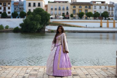 15. yüzyıl elbisesi giyen uzun kıvırcık saçlı güzel Latin kadın İspanya 'daki Guadalquivir nehrinin kıyısında duruyor. Arka planda şehrin diğer tarafını görebilirsiniz.